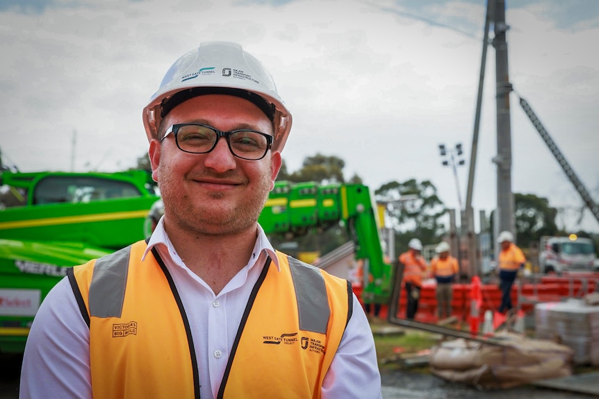 A man wearing a hard hat and hi vis smiling at the camera with big machinery in the background.
