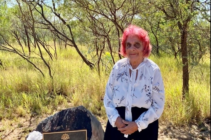An elderly woman with pink hair stands at a war memorial in the bush