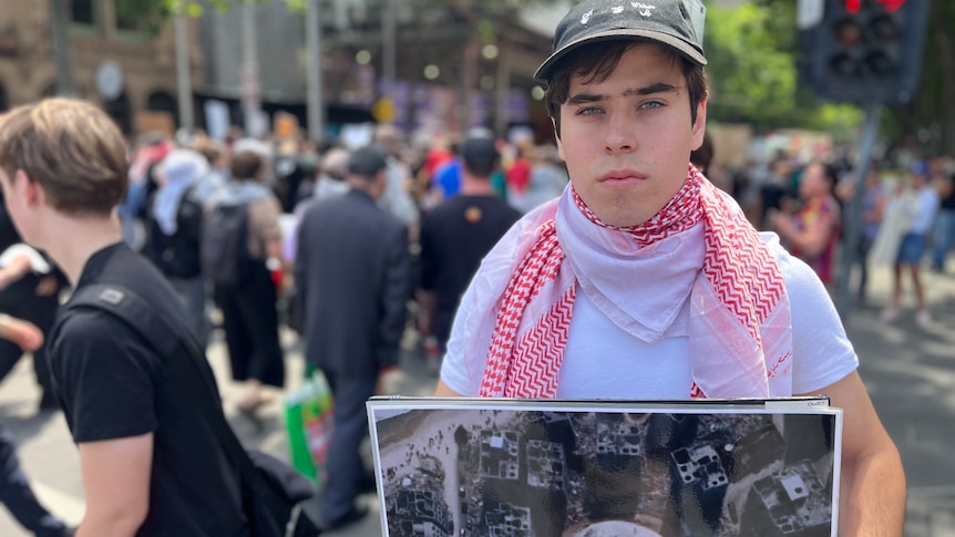 Teenage boy in a white top and black cap holding up a photo in the middle of a protest.