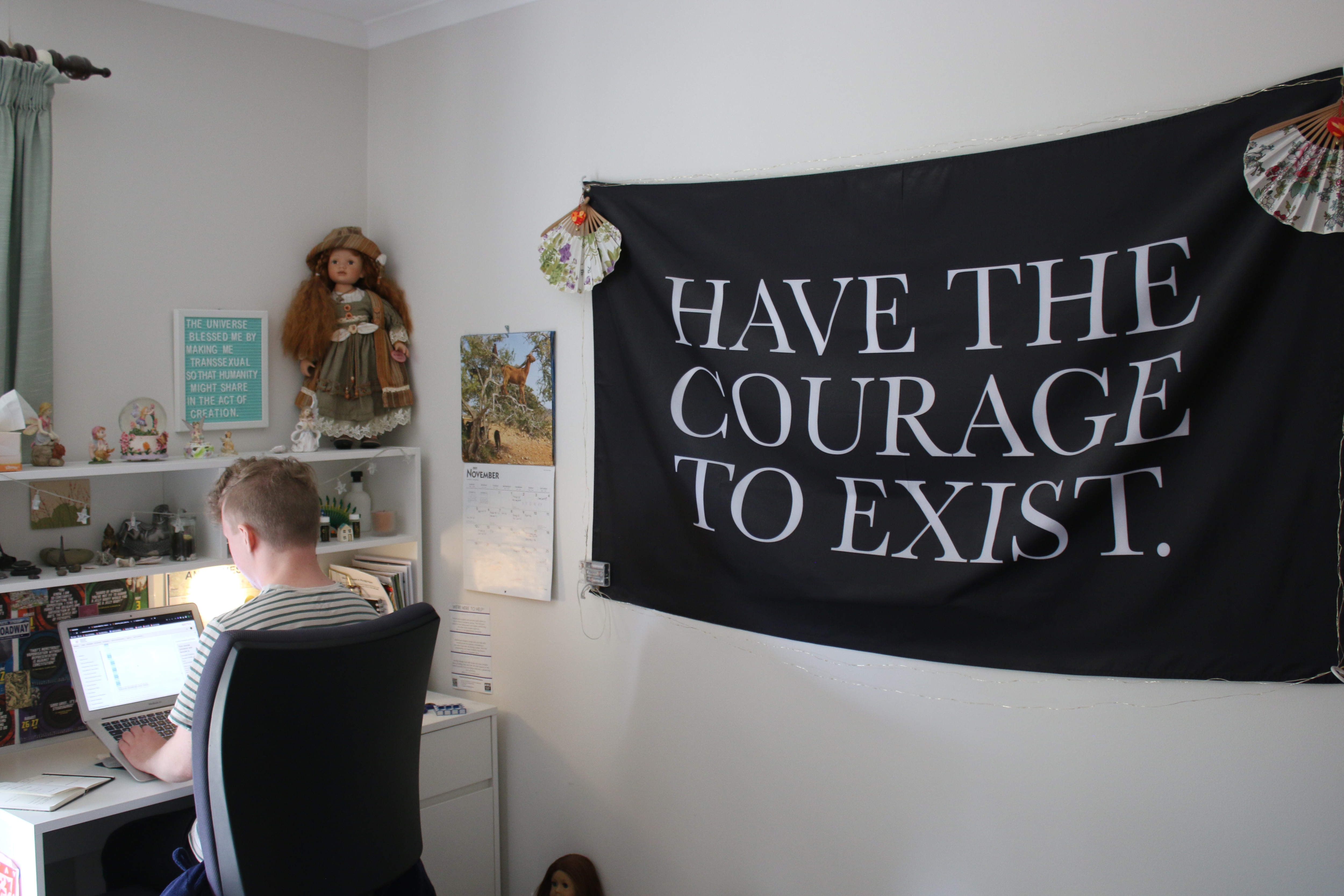 A young man sits at a computer desk in a room with a big banner on the wall that says "Have The Courage To Exist".