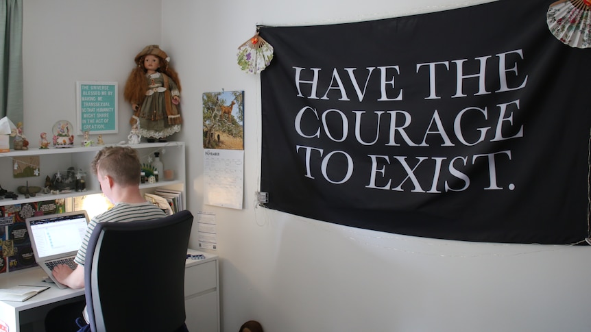 A young man sits at a computer desk in a room with a big banner on the wall that says "Have The Courage To Exist".