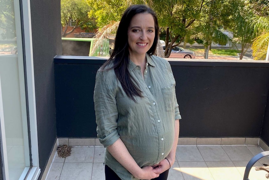 A pregnant woman with dark hair smiles on a balcony.