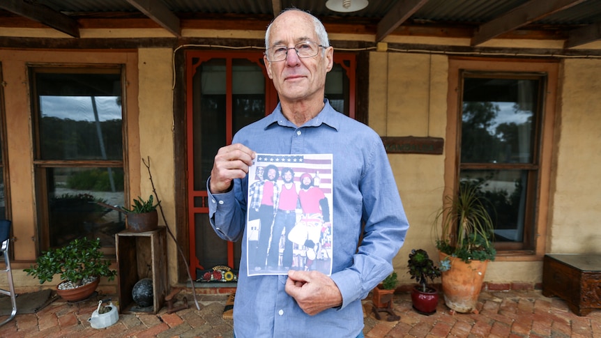 Bill Wiglesworth outside his house holding a picture of himself and two friends at an American Independence Day party.