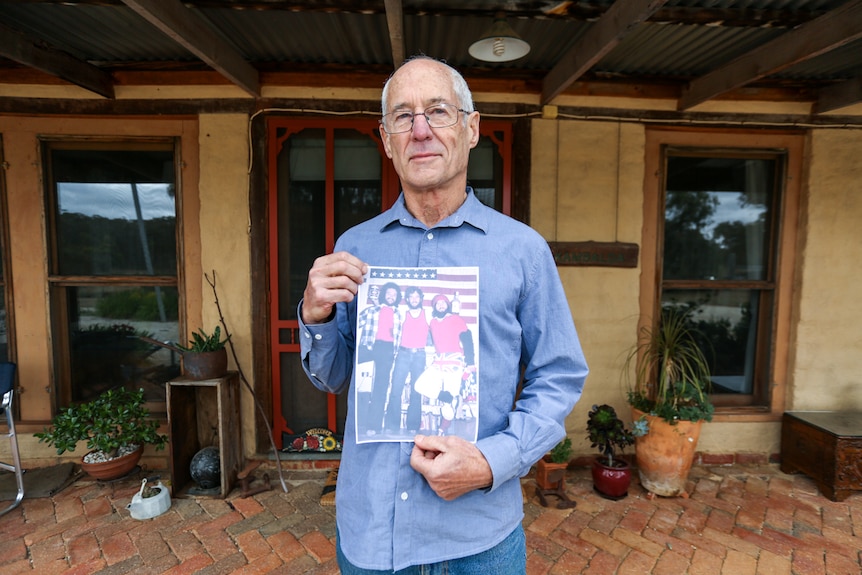 Bill Wiglesworth outside his house holding a picture of himself and two friends at an American Independence Day party.