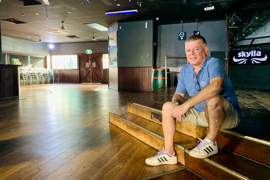 A man in a blue shirt and sneakers sits on the edge of an empty dancefloor.