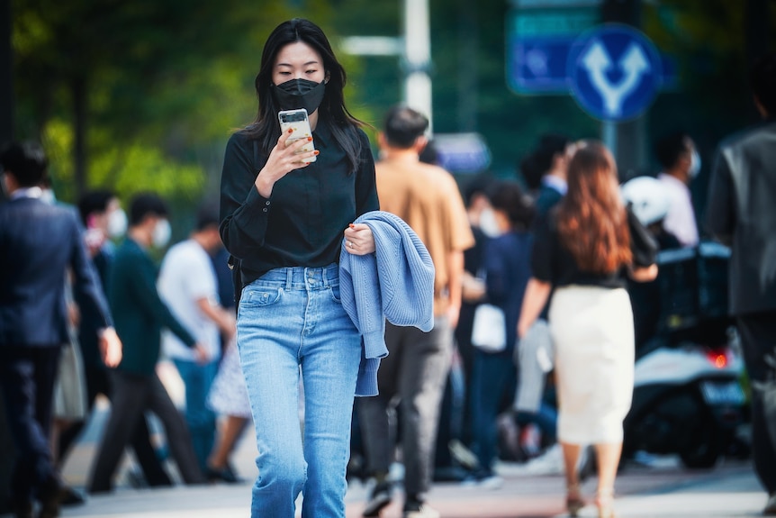 A woman on a street wearing jeans, a black top and a mask looks at her phone.