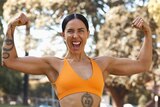 A woman in an orange exercise top flexes with both arms and smiles at the camera with a tattoo on her torso