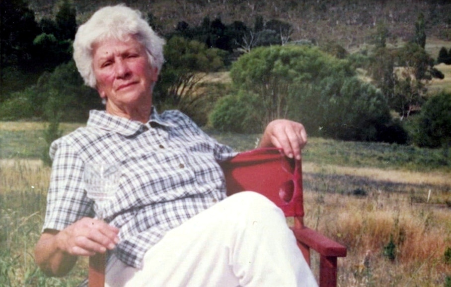 An older woman with short grey hair reclines on a red camping chair in an unidentified paddock.