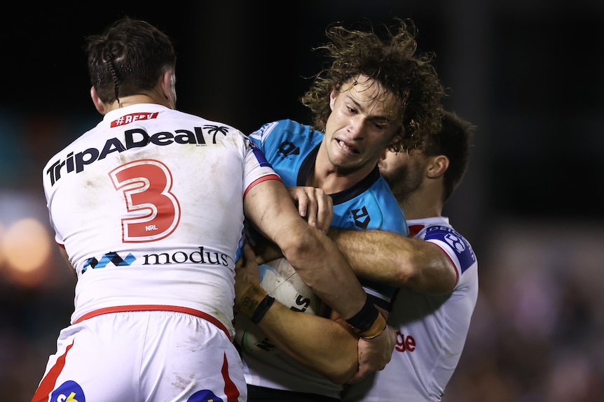 A Cronulla NRL player holds the ball as he is tackled by two St George Illawarra opponents.