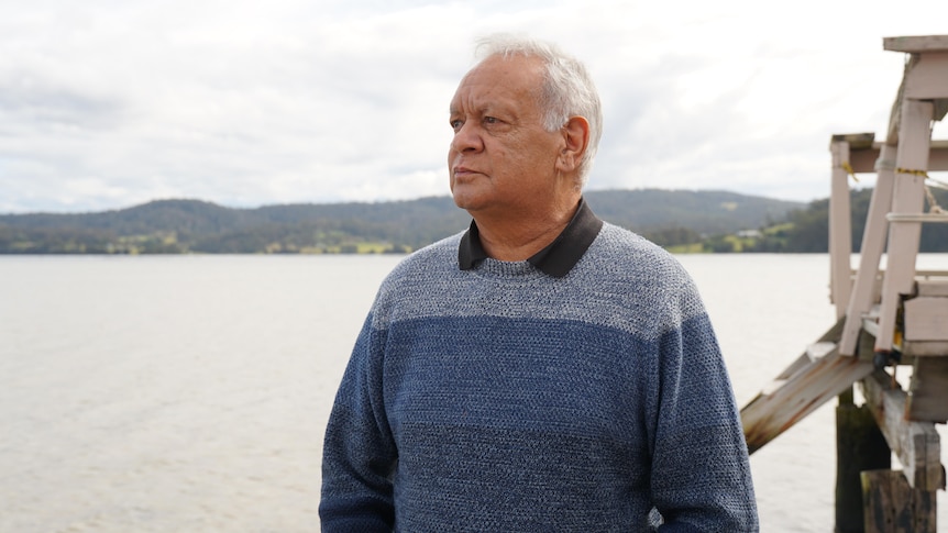 An older Aboriginal man with receding grey hair stands by the water, looking out into the lake pensively.