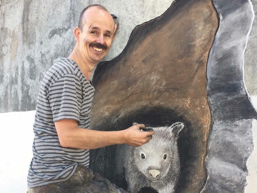 Man stands next to mural of a wombat on the side of a bridge. 