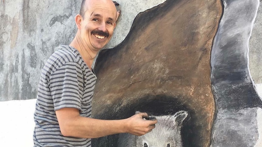 Man stands next to mural of a wombat on the side of a bridge. 