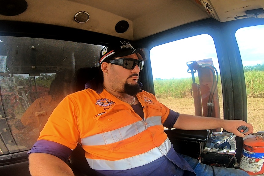 a man in a high vis work shirt inside the cab of a harvester he's operating in a cane field