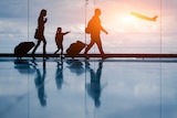 Silhouettes of a family walk past an airport window as a plane takes off outside.