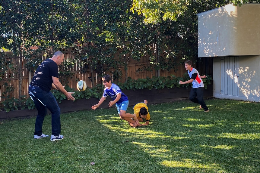 A man plays rugby in the backyard with two young teenage boys.