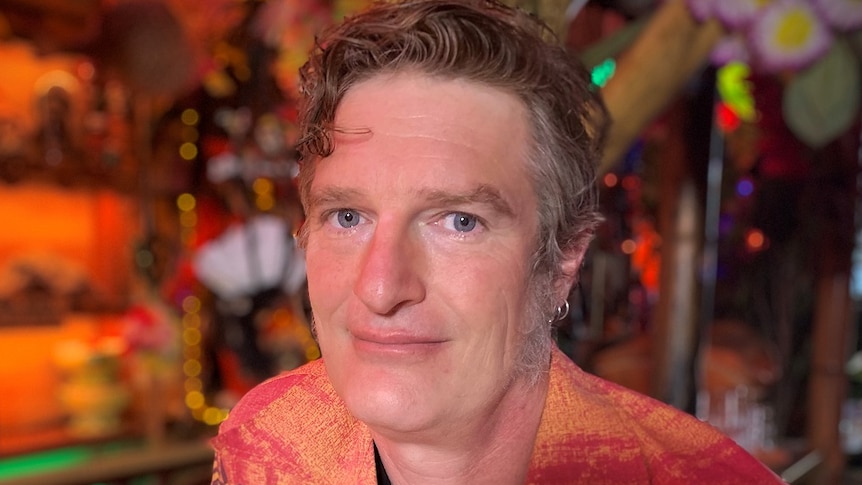 Josh Collins stands in front of a bar, dressed in a colourful patterned shirt.