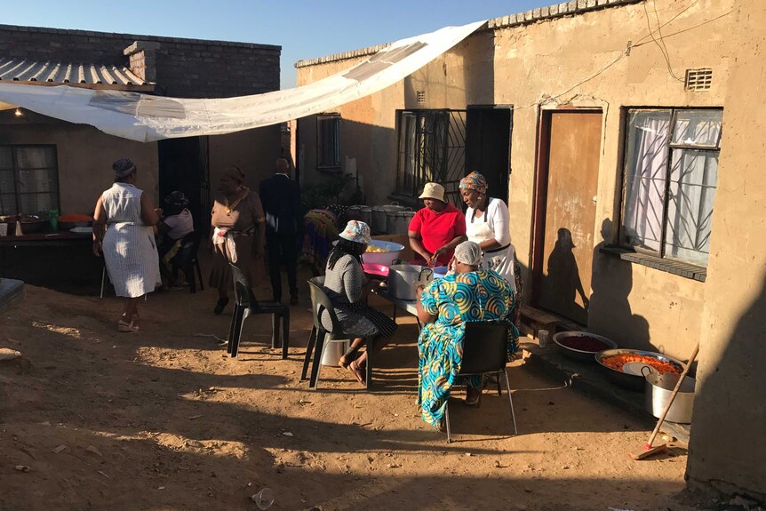 Women gather at a table outside a small house, preparing food.