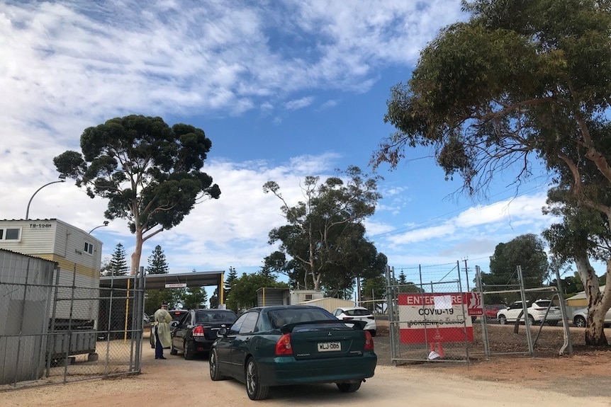 Cars outside lined up for a COVID-19 drive-through test, with blue sky in background and brown dirt in foreground.