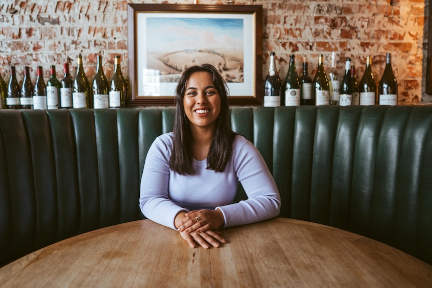 A woman sits in a grey dress and smiles. She is in a booth with wine bottles lined up behind her.