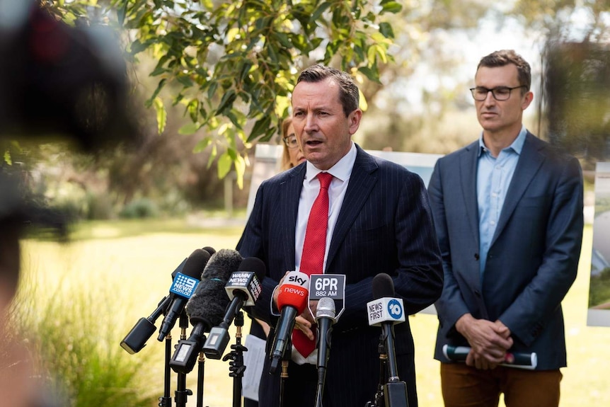 Mark McGowan wearing a pin stripe suit with a red tie, standing at a podium with microphones with two politicians behind.
