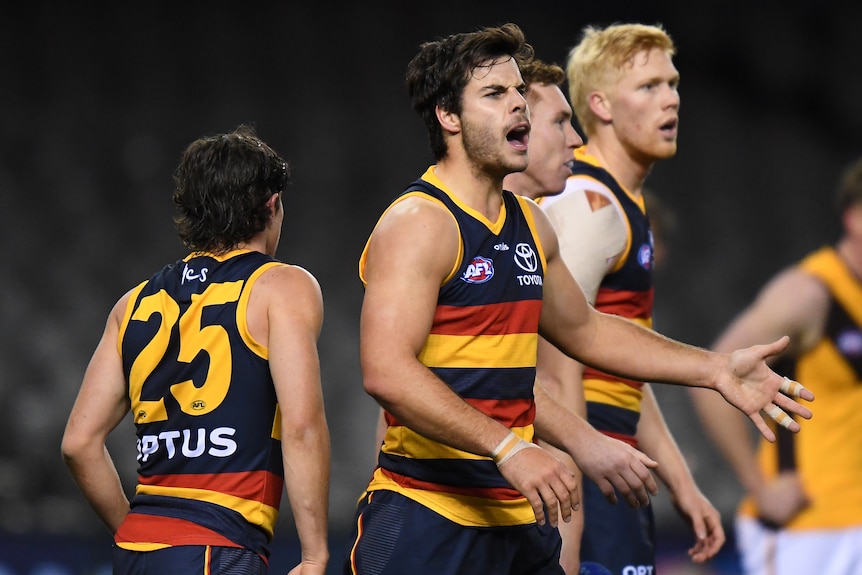 An AFL player in black, red and yellow with a beard shouts in joy surrounded by teammates. 