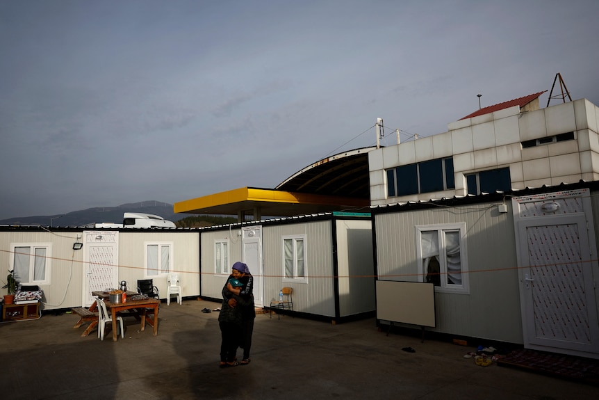 Two people embrance outside a row of white container homes.