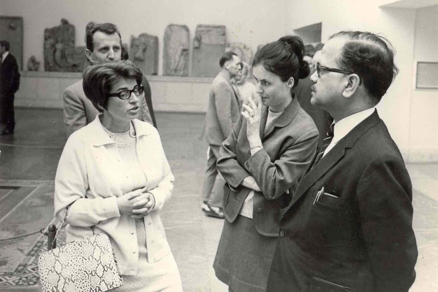 A black and white photo with a two women and a man standing in a museum. One woman is talking.