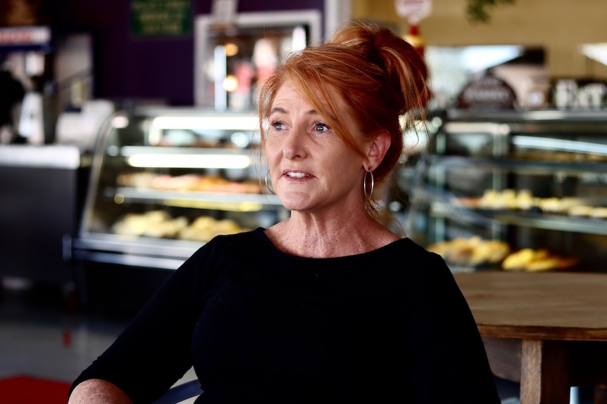 A lady in a black top looks of camera, with counters of baked goods behind her
