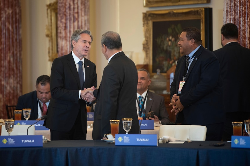 Two suited men shake hands amid a formal meeting, while other watch and smile.