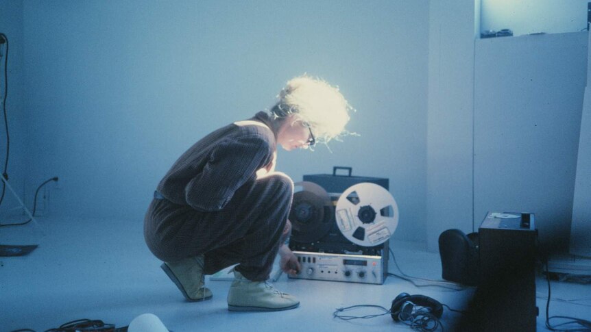 A woman in a blue room crouches down in front of a tape machine.