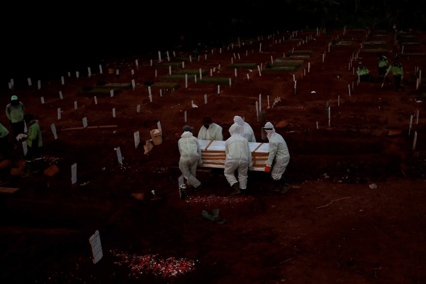 Five men wearing hazmat suits carry a coffin among a sea of freshly dug graves as grave diggers dig more graves.