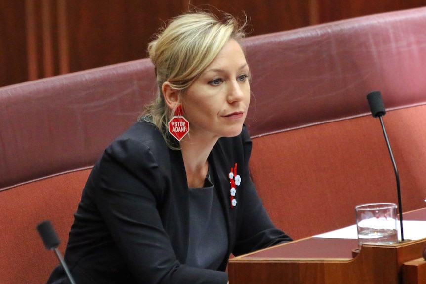 Larissa Waters sits in a red seat in the Senate. She is wearing red earrings in the shape of stop signs that say STOP ADANI.