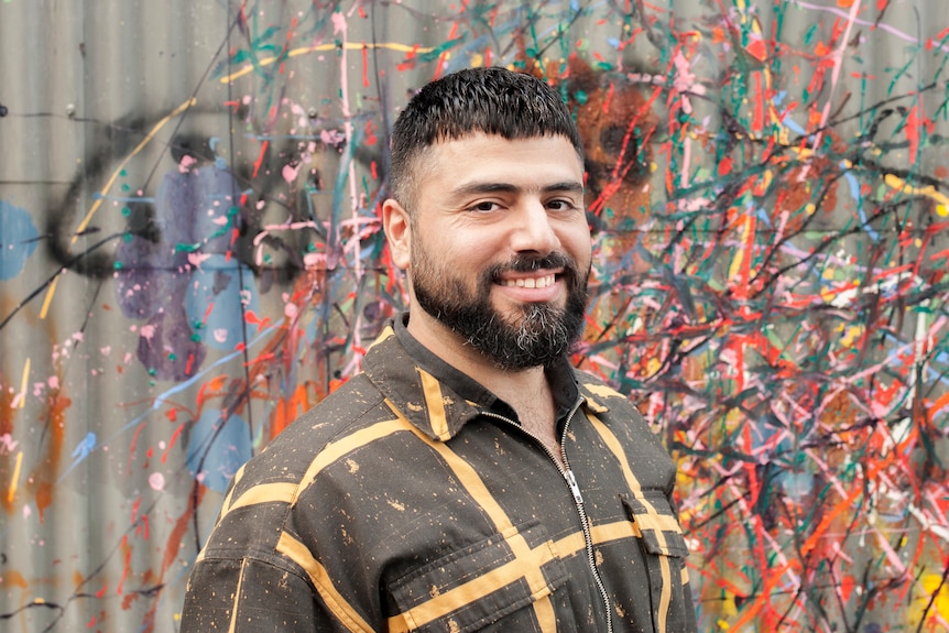 A smiling man with a short beard and hair stands in a front a metal fence with colourful paint splattered across it