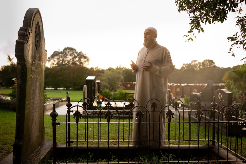 A man wearing a traditional Sunni Muslim baya prays at a grave site as the sun sets behind him.