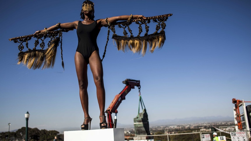 An African woman dressed as a bird stands on a plinth arms outstretched, a statue of man is removed by a crane in the background