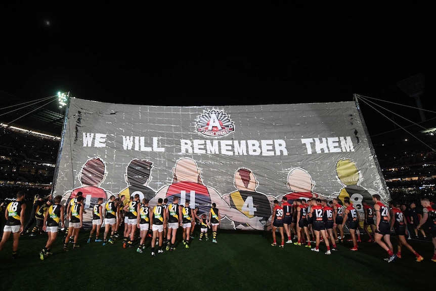 Richmond and Melbourne players walk through banner saying 'we will remember them'
