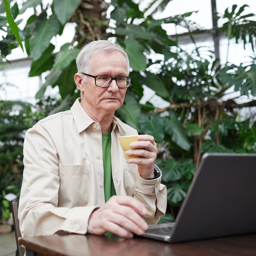 An older man holding a coffee cup stares morosely at a laptop has he types in a greenhouse cafe