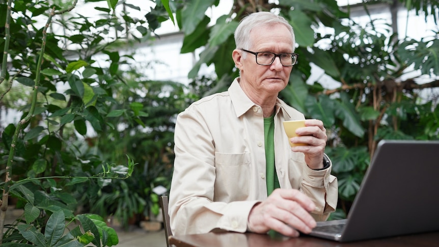 An older man holding a coffee cup stares morosely at a laptop has he types in a greenhouse cafe