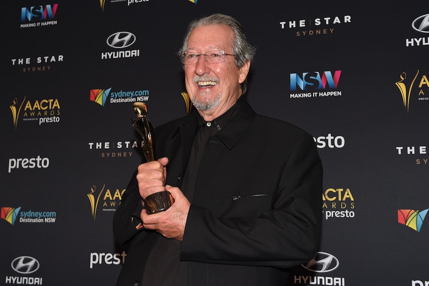 Michael Caton smiles holding an AACTA award standing in front of a promotional board