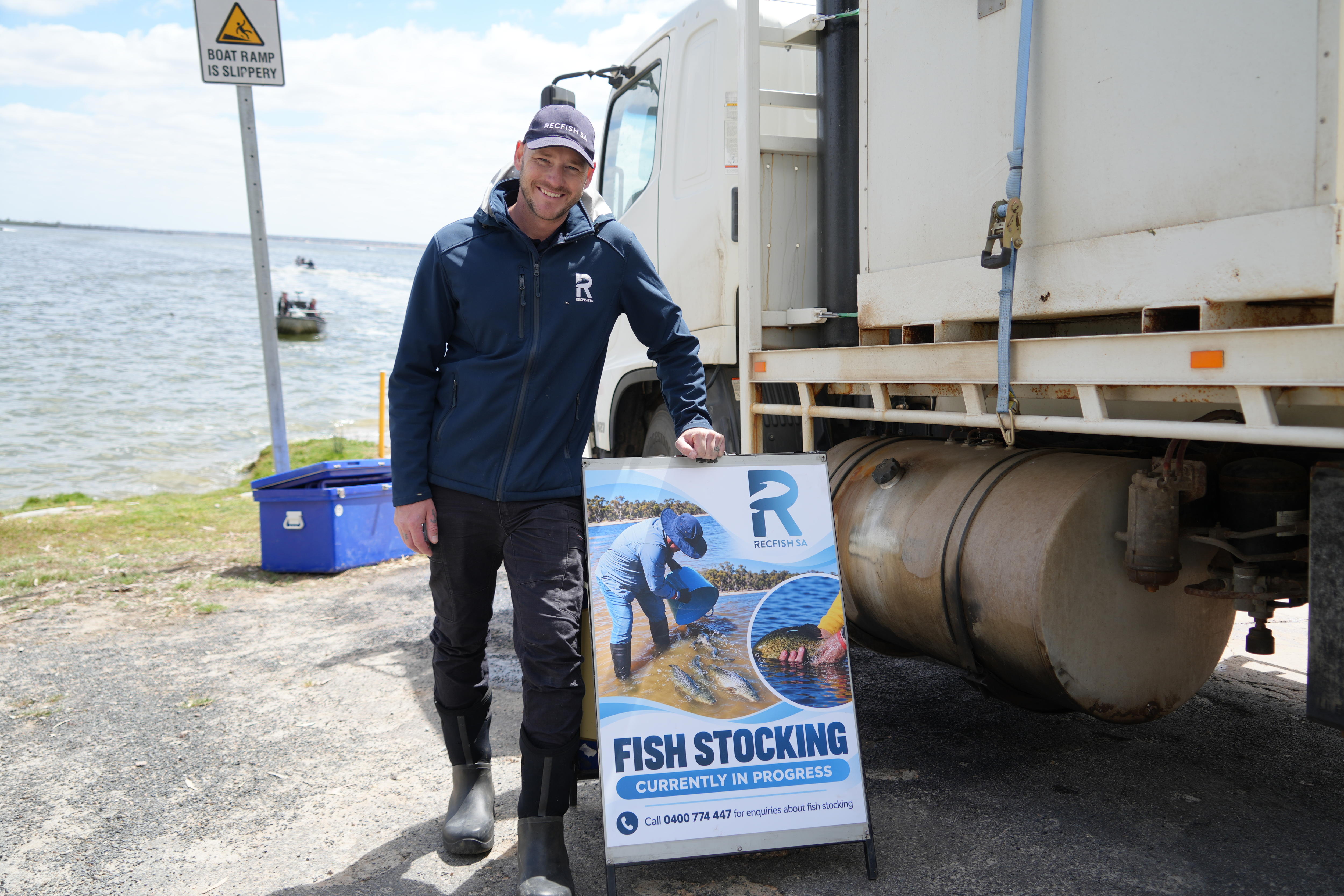 A smiling man wearing a cap, dark clothes and gum boots next to a sign and large truck on the shores of a large lake.