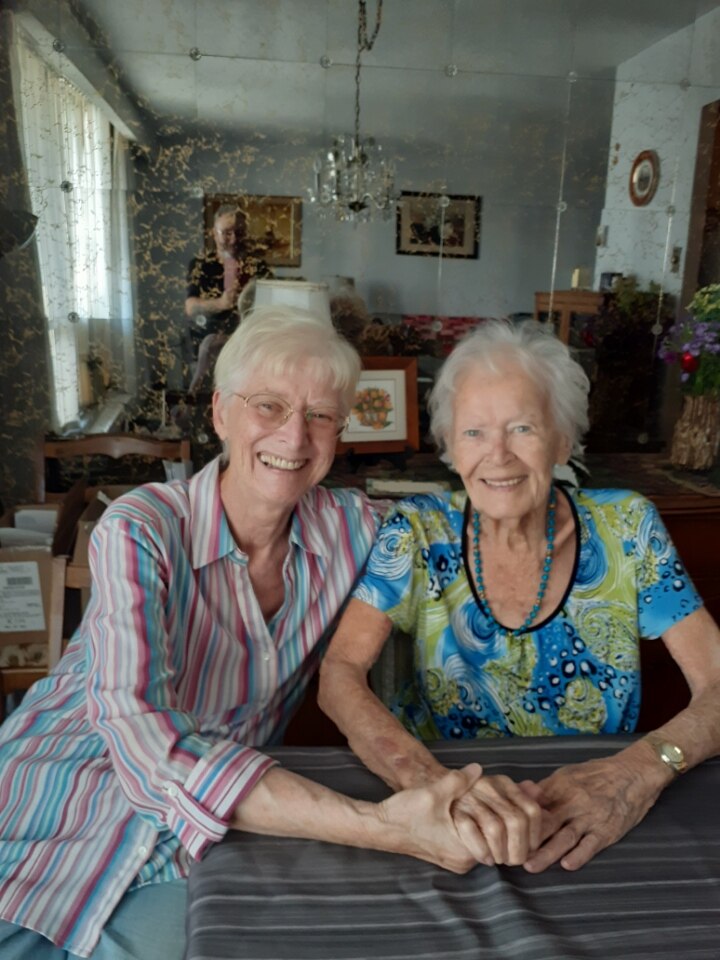 Two women sit at a dining table, holding hands and smiling at camera.