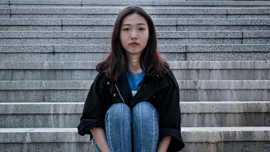 A young woman with long brown-red hair, looking intently to camera, sitting on concrete steps.