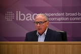 A man sits behind a desk, in front of a purple IBAC sign.