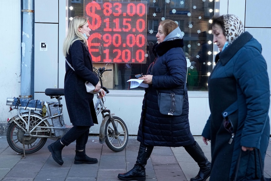 Two older women with headscarves and one younger woman pushing a bike walk past a window with an LED currency exchange rate sign