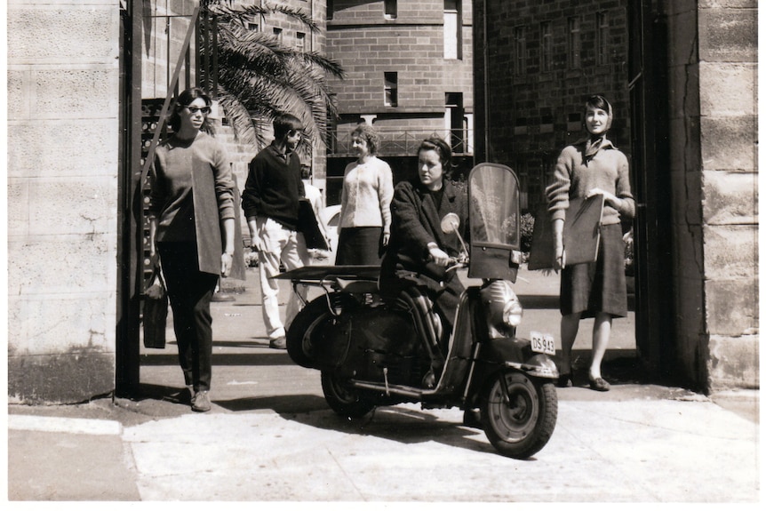 A group of students are posed at the Entry to East Sydney Technical College. At the centre, Vivienne Binns rides a scooter.