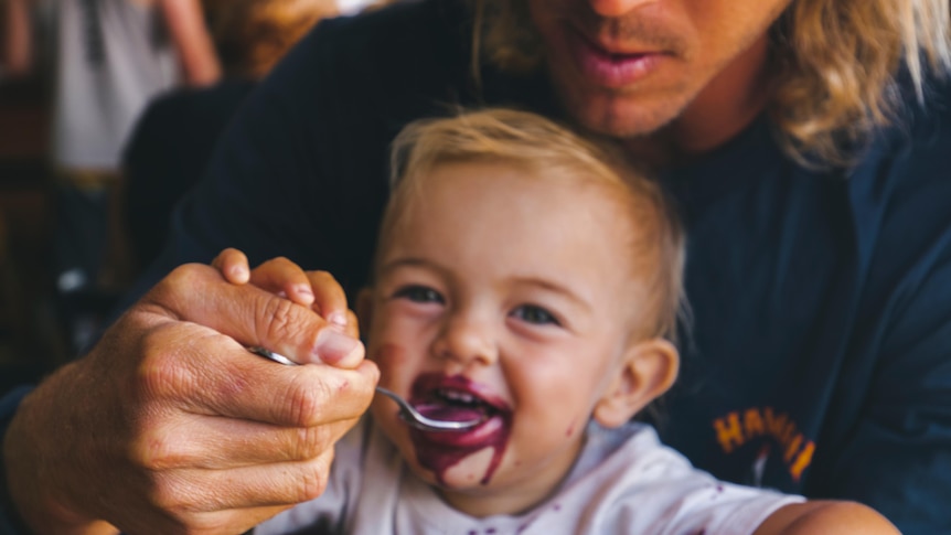 Close-up photo of just-visible man holding and feeding a baby with a spoon. The baby smiles with purple food around its mouth.