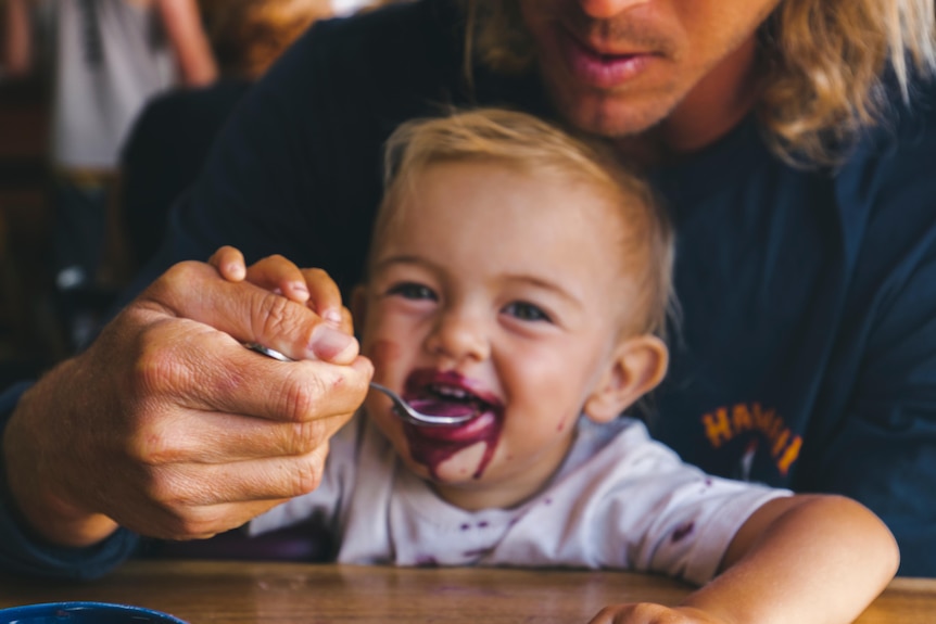 Close-up photo of just-visible man holding and feeding a baby with a spoon. The baby smiles with purple food around its mouth.