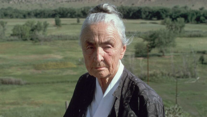 An older woman sitting in front of grassy plains and mountains in the distance.