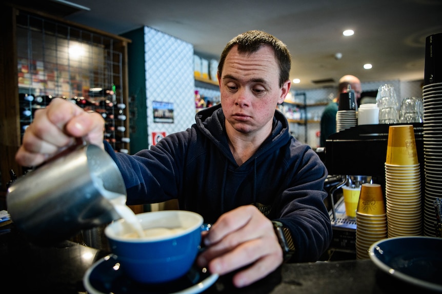 A white man with Down syndrome pouring milk into a coffee at an espresso machine