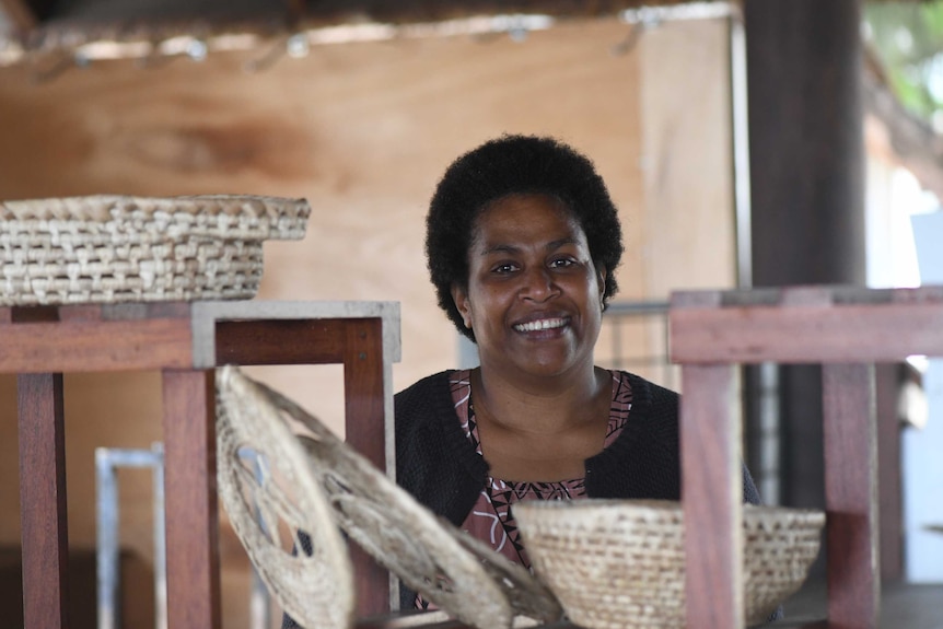 Joslyn Garae Lulu stands behind shelves with some of the woven goods she sells. She is smiling for the camera.